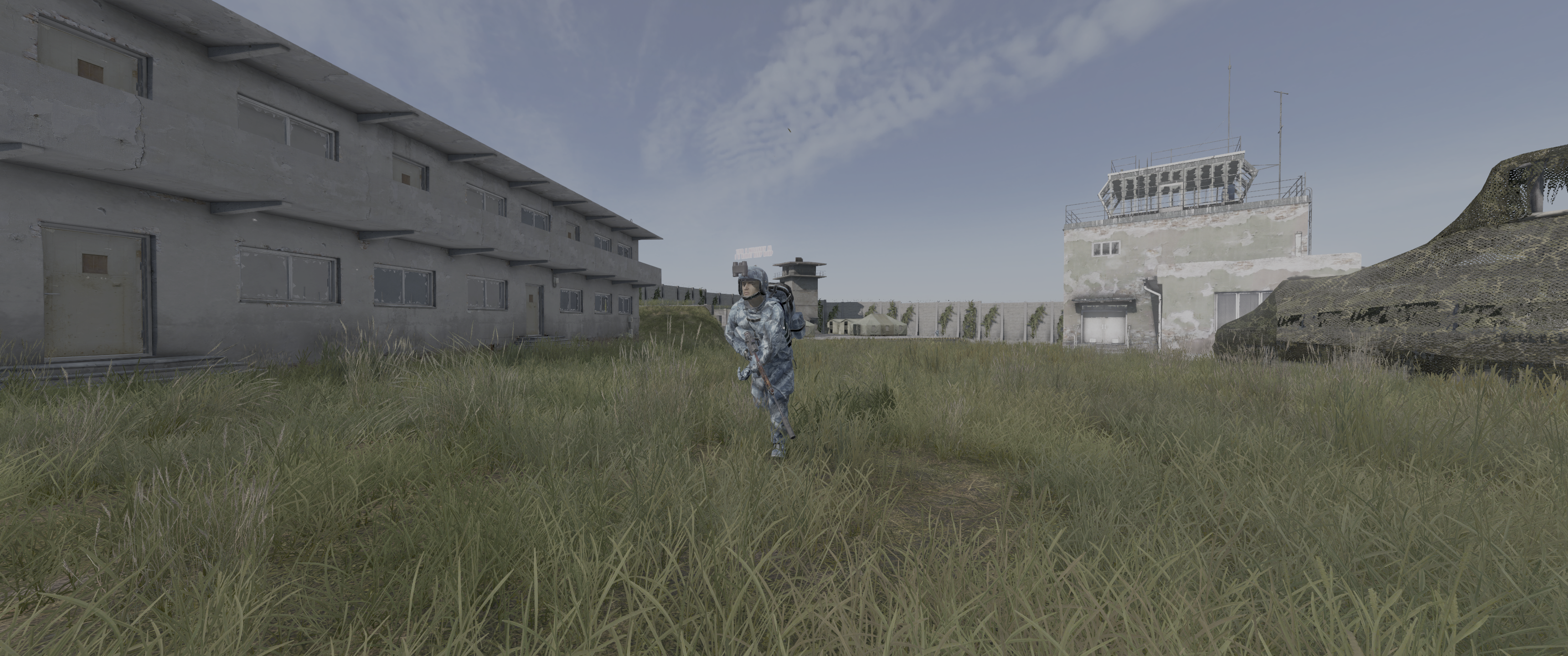 A soldier in camouflage clothing walks through tall grass in an abandoned military compound with two buildings in the background.