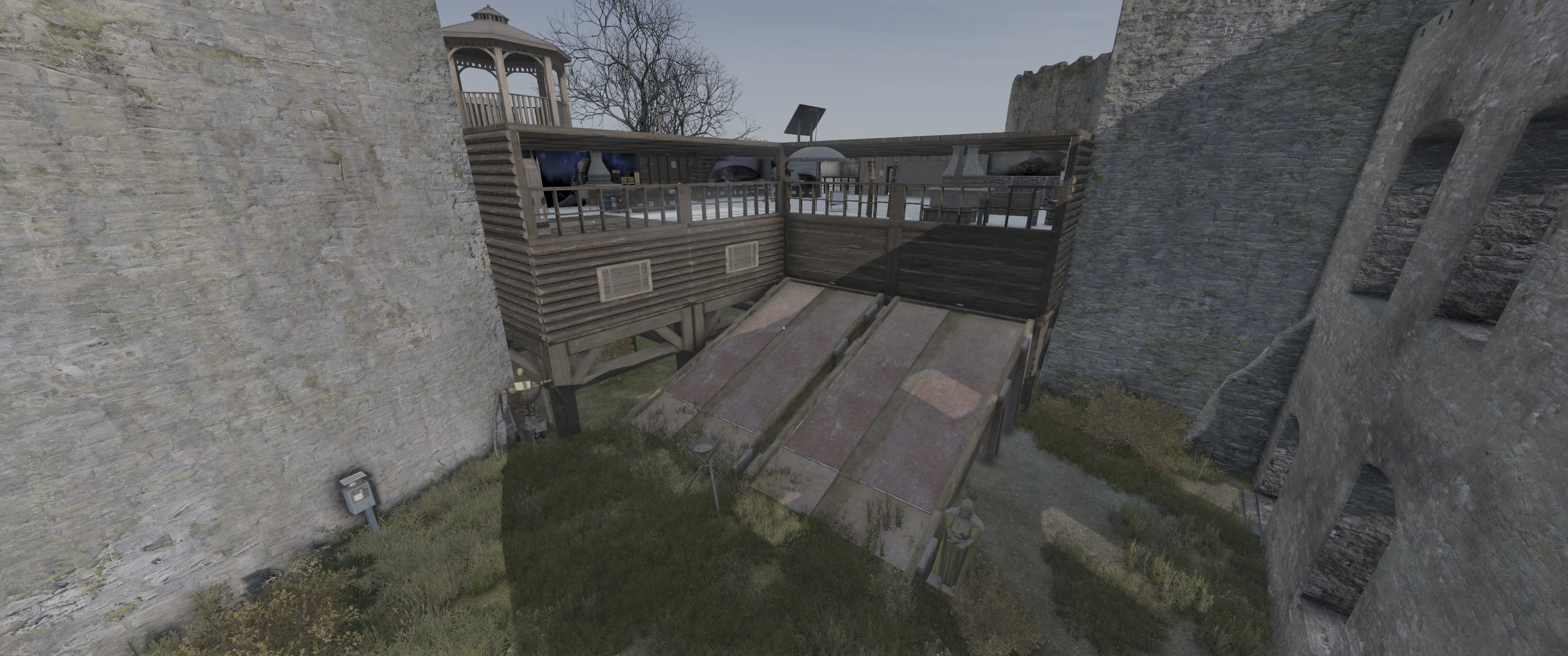 A wooden structure with a balcony on an elevated platform, positioned near a stone wall. The structure features a gazebo-like roof and has a ramp leading down to grass-covered ground.